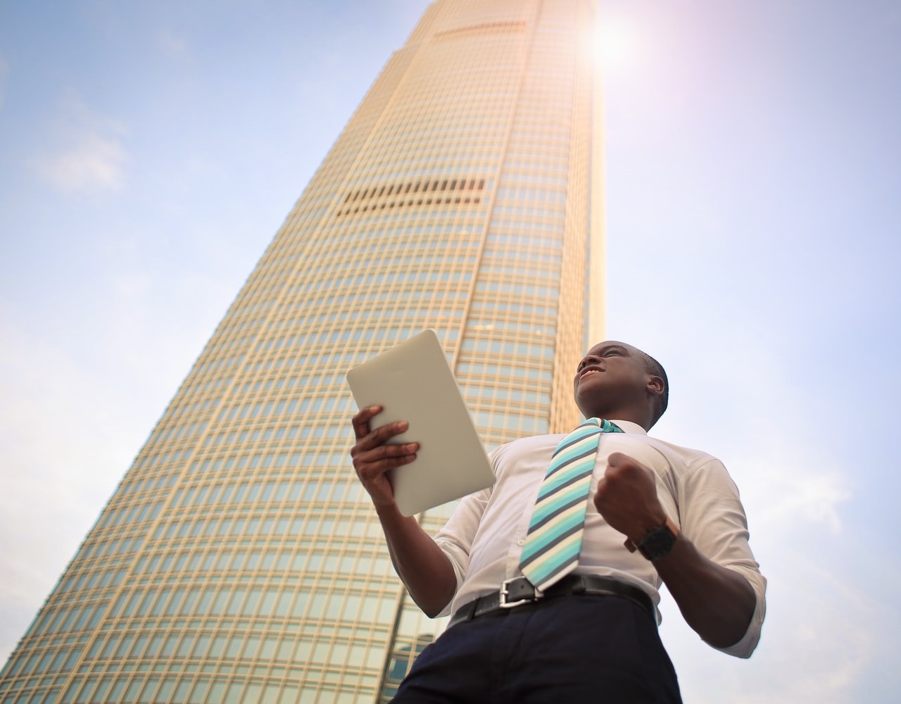 Photo of a tidily dressed young man holding documents, with the background of a tall building and a blue, sunny sky
